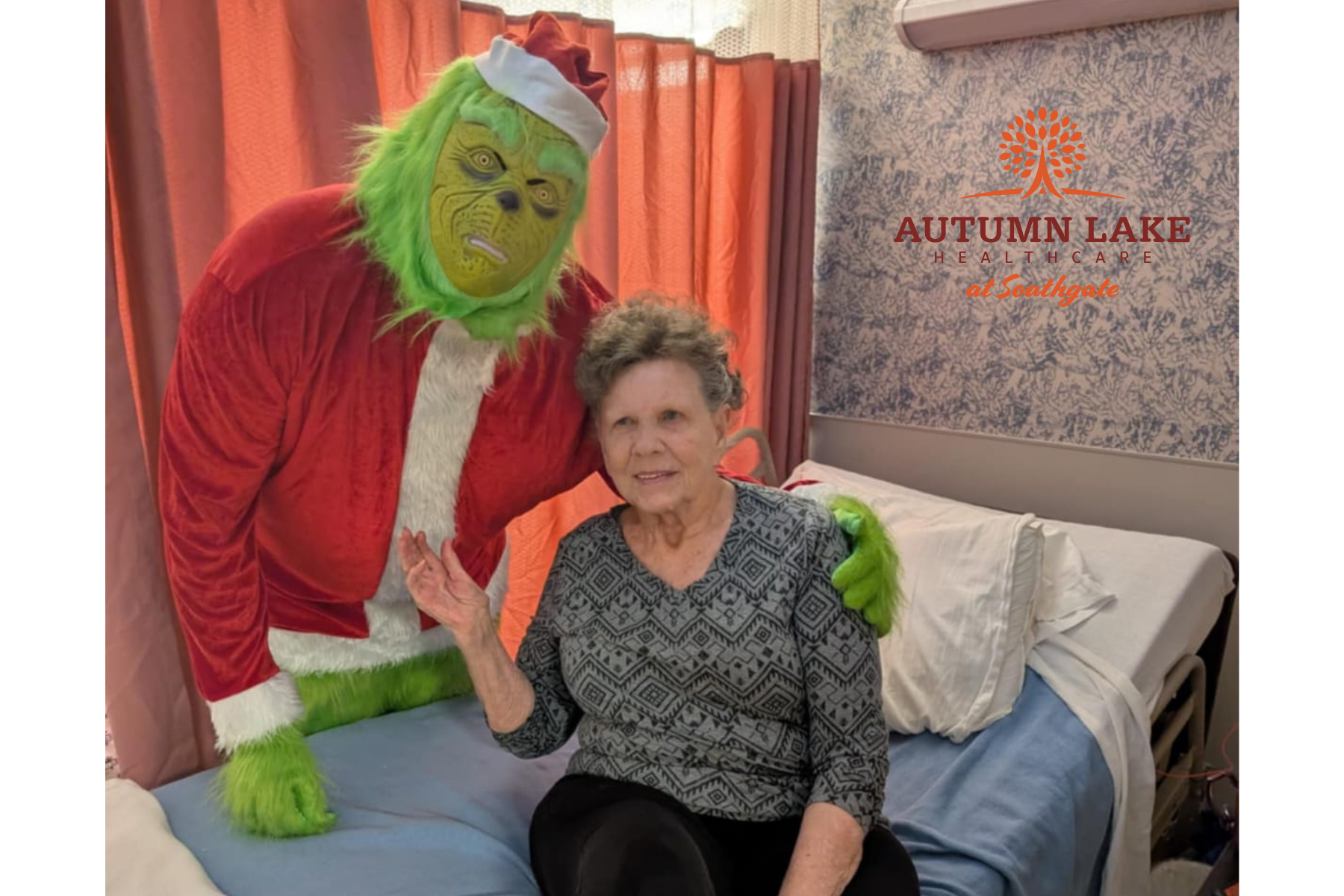 A nursing home resident sits on her bed and poses for a photo with a person dressed in a Grinch costume during the holidays.